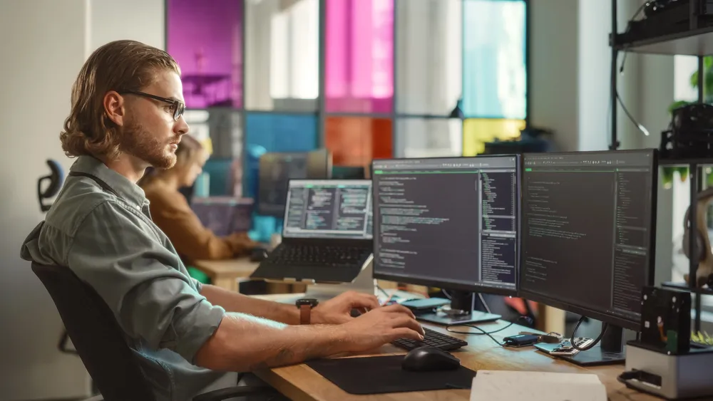 Web developer piše kod na dva monitora u modernom uredu